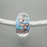 Decorative faceted glass bead with floral design and aqua core on a silver bangle against a grey background