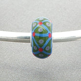 Decorative opaque bead with green and blue pattern on a silver bangle against a grey background