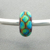 Decorative glass bead with blue and green pattern on a silver bangle against a grey background