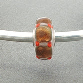 Glass bead with golden squares and orange accents on a silver bangle against a grey background