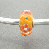 Glass bead with an orange and yellow floral design on a silver bangle against a grey background