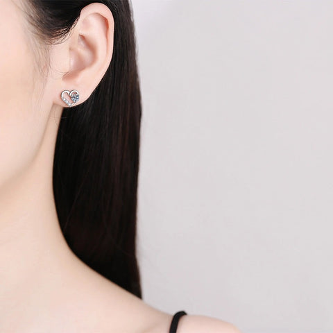 Close-up of a person wearing a heart-shaped earring with a clear gemstone on a plain background