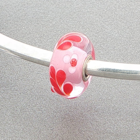 Glass bead with pink core and red and white leaf pattern on a silver bangle against a grey background