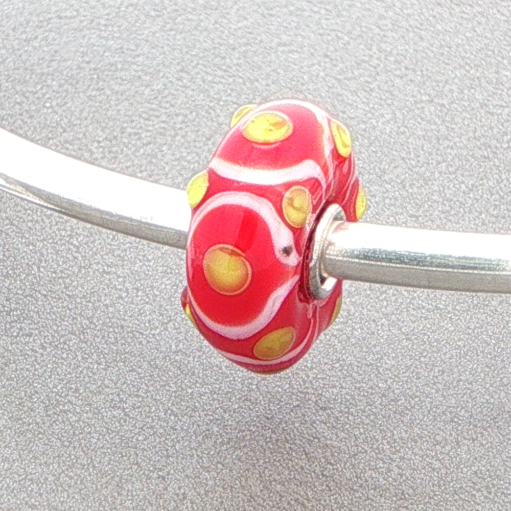 Red and white glass bead with tactile yellow dots on a silver bangle against a grey background