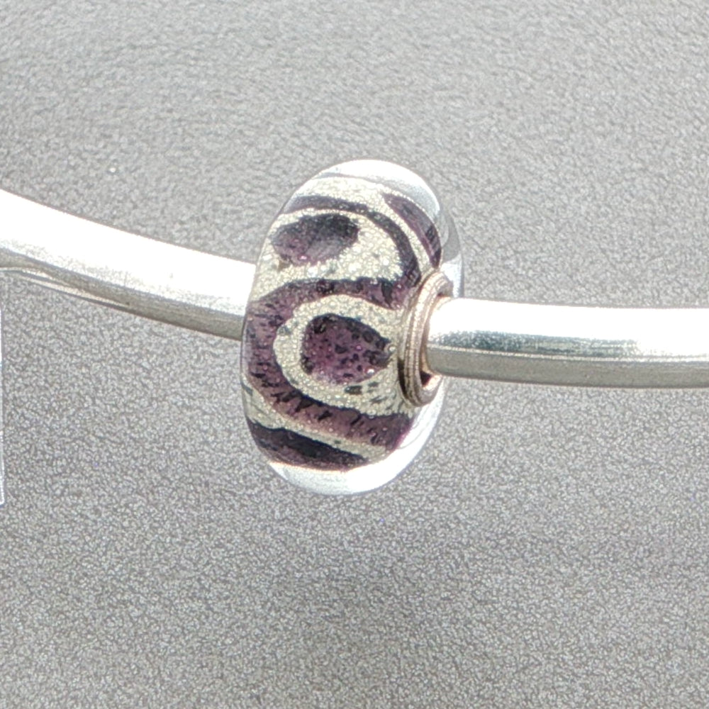 Glass bead with purple and silver glitter design on a silver bangle against a grey background