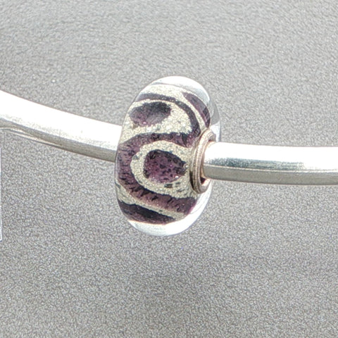 Glass bead with purple and silver glitter design on a silver bangle against a grey background