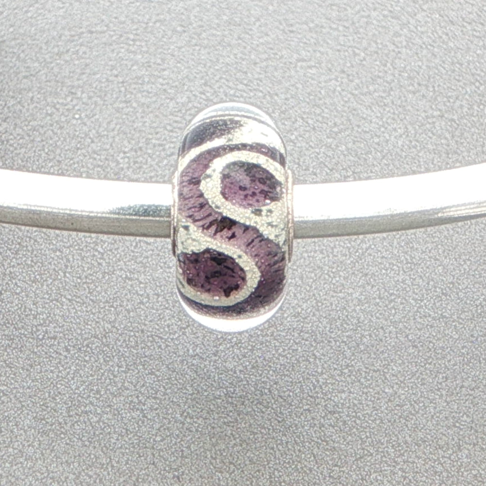 Murano glass bead with purple and glitter pattern on a silver bangle against a grey background