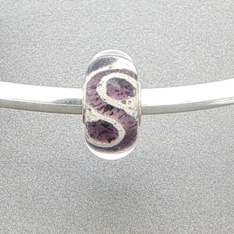 Murano glass bead with purple and glitter pattern on a silver bangle against a grey background