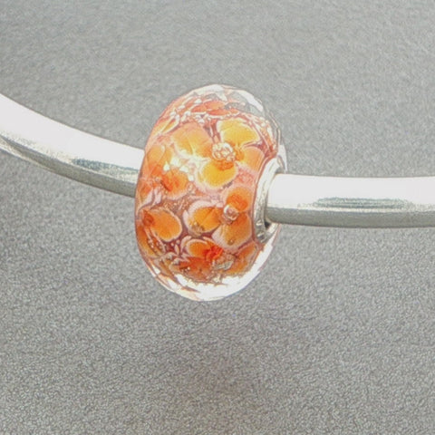 Faceted glass bead with orange floral pattern on a silver bangle against a grey background