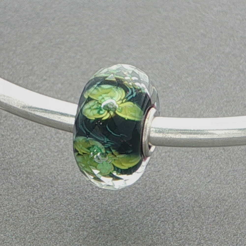 Faceted glass bead with green core, green floral design and sprinkle of gold glitter on a grey background