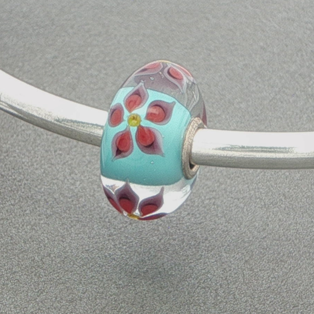 Glass bead with floral design and aqua core on a silver bangle against a grey background