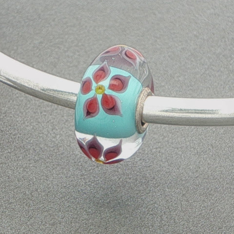 Glass bead with floral design and aqua core on a silver bangle against a grey background
