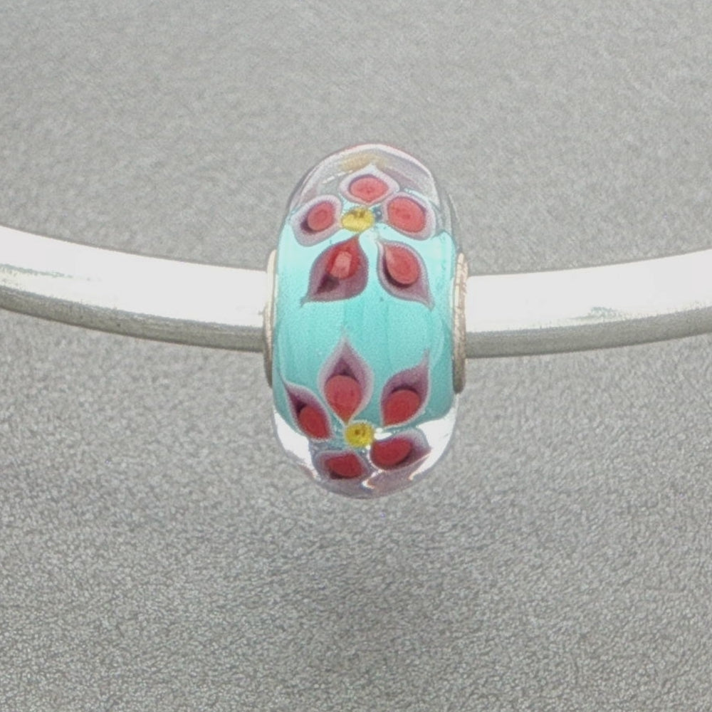 Decorative glass bead with floral detail and aqua core on a silver bangle against a grey background