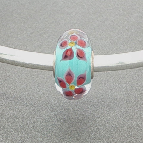 Decorative glass bead with floral detail and aqua core on a silver bangle against a grey background