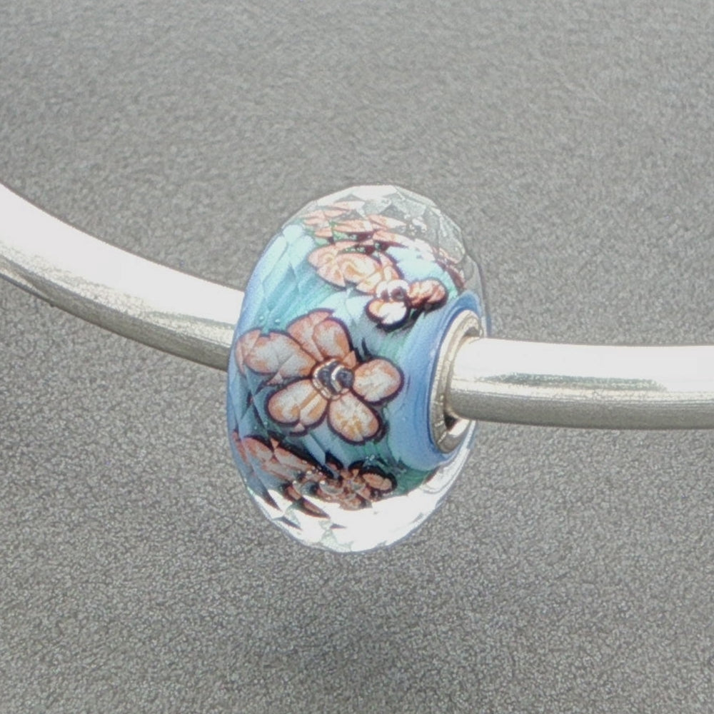 Decorative faceted glass bead with an aqua core and floral design on a silver bangle against a grey background