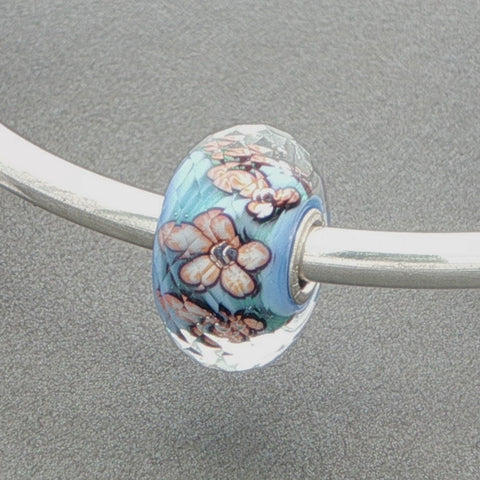 Decorative faceted glass bead with an aqua core and floral design on a silver bangle against a grey background