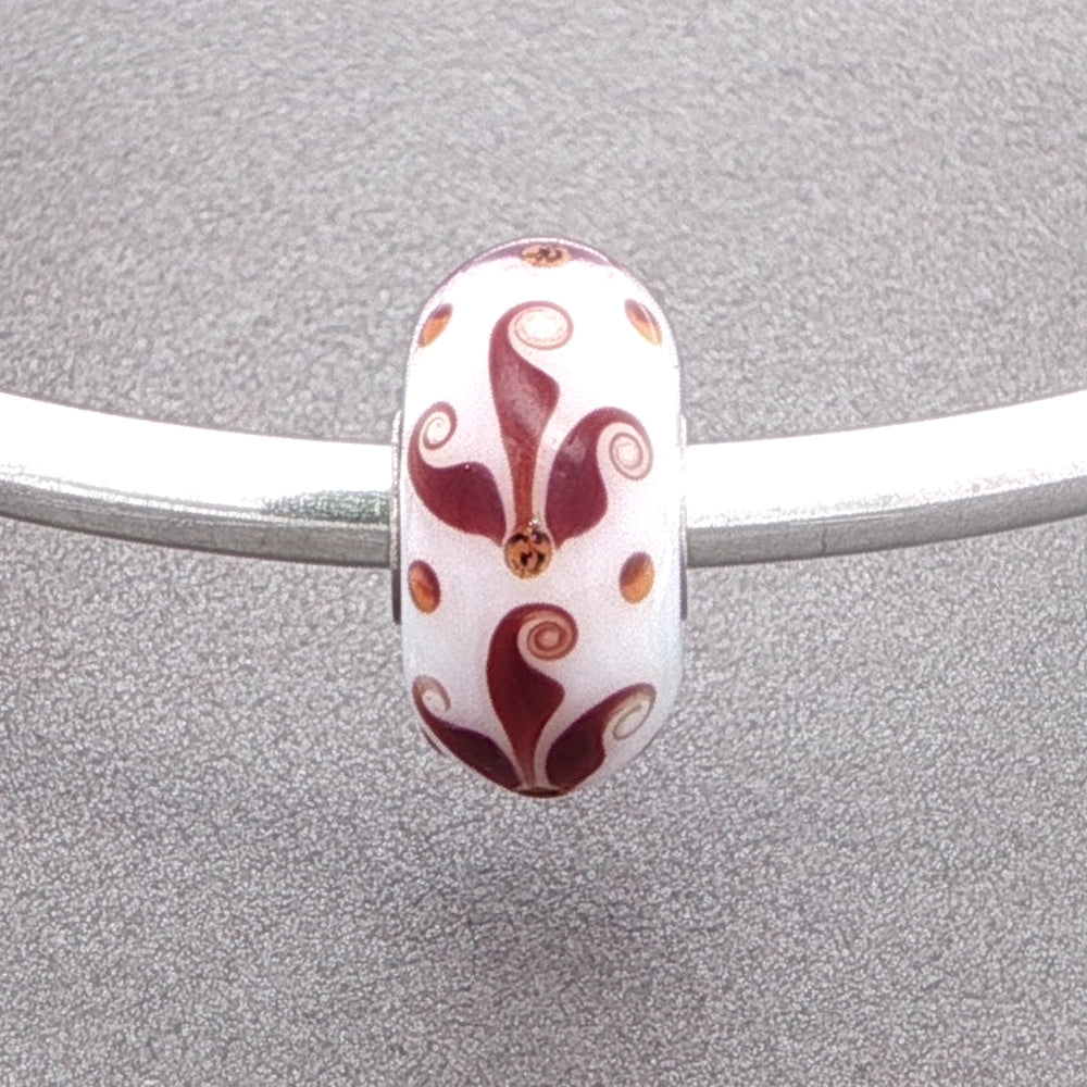Decorative opaque white glass bead with a brown flourish type pattern on a silver bangle against a grey background