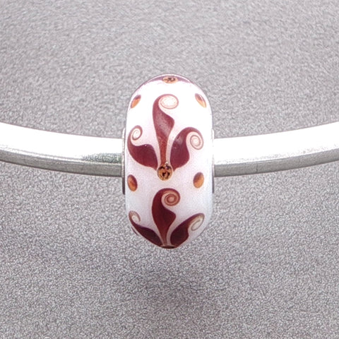 Decorative opaque white glass bead with a brown flourish type pattern on a silver bangle against a grey background
