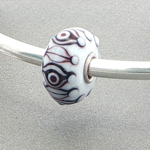 White opaque bead with a eye pattern in black and brown on a silver bangle against a grey background