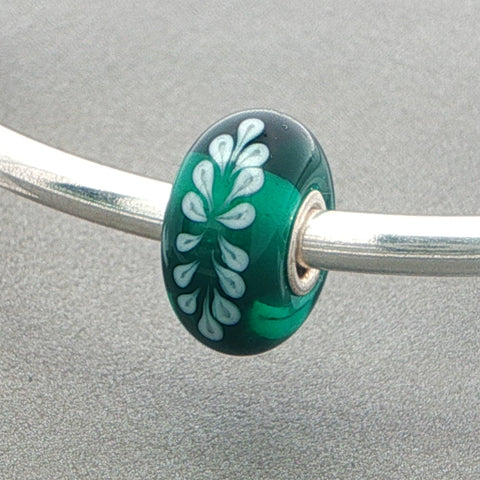Green glass bead with a white fern-like pattern on a silver bangle against a grey background