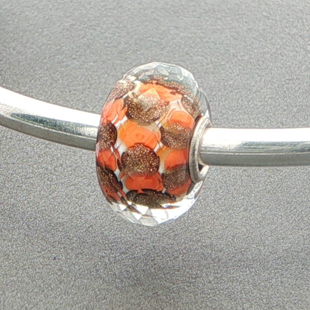 Faceted glass bead charm with orange and brown pattern on a silver bangle against a grey background