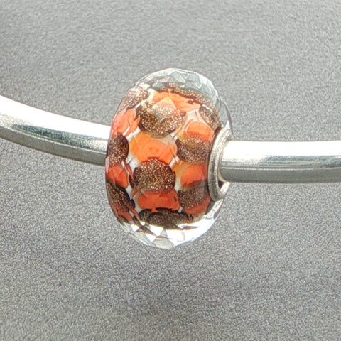 Faceted glass bead charm with orange and brown pattern on a silver bangle against a grey background