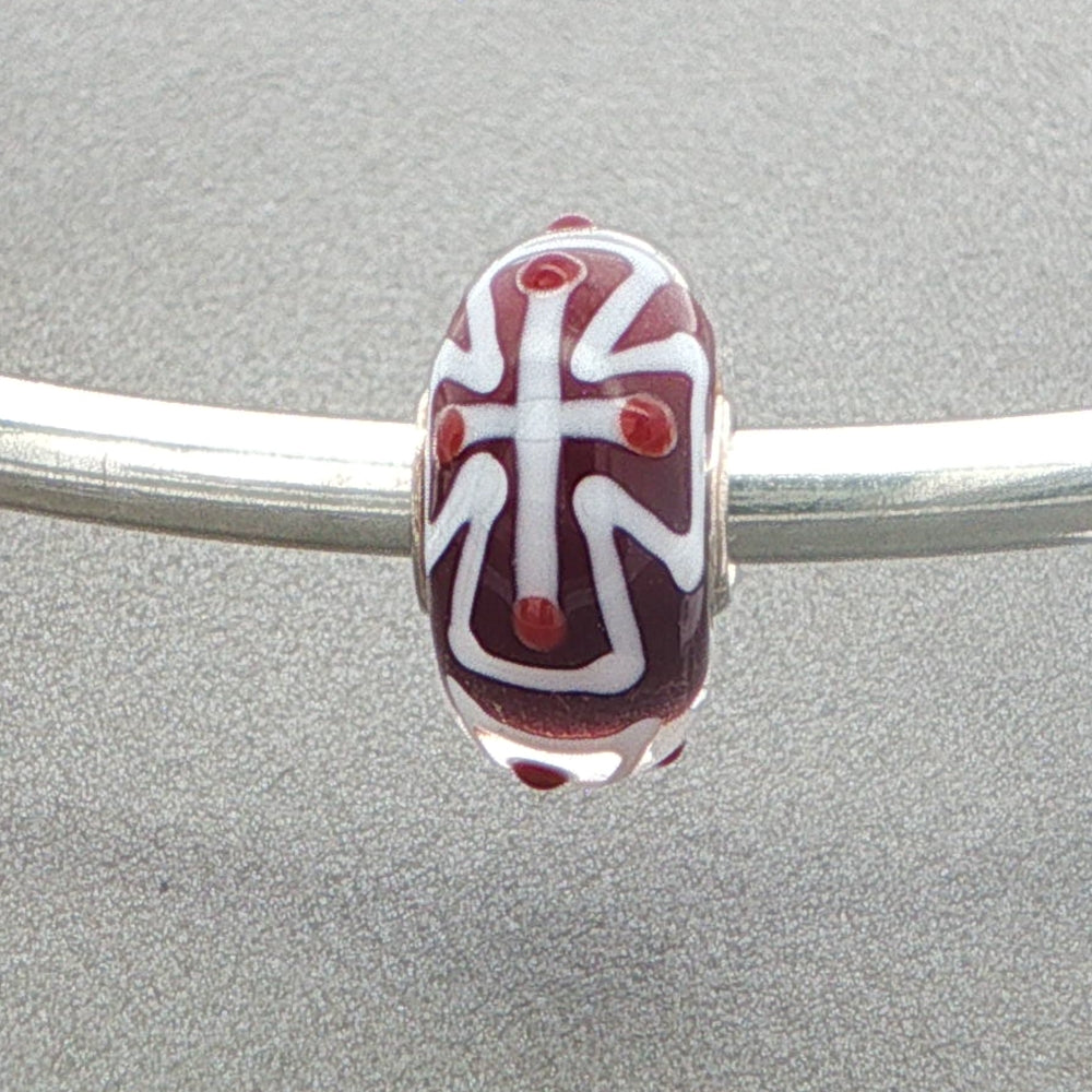 Glass bead with a white cross design on a silver bangle against a grey background