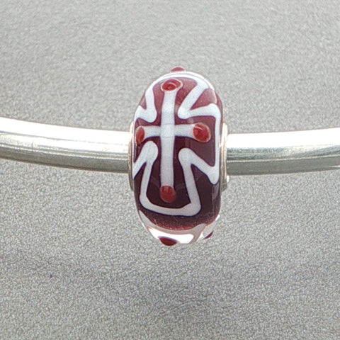 Glass bead with a white cross design on a silver bangle against a grey background