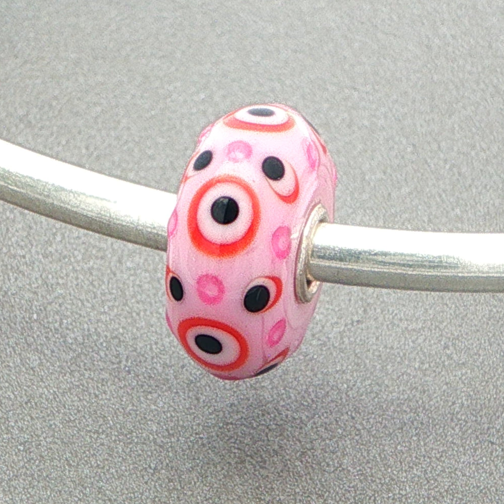 Pink opaque glass bead with pink and black dots with red circles on a silver bangle against a grey background
