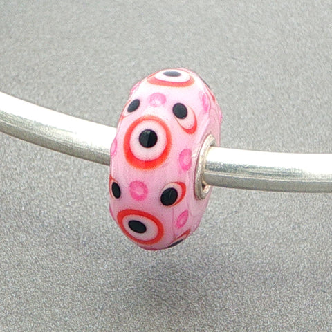 Pink opaque glass bead with pink and black dots with red circles on a silver bangle against a grey background