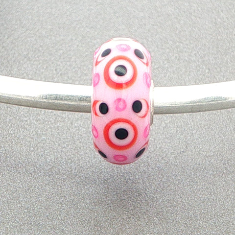 Murano glass bead with pink opaque core with pink and black dots with red circles on a silver bangle against a grey background