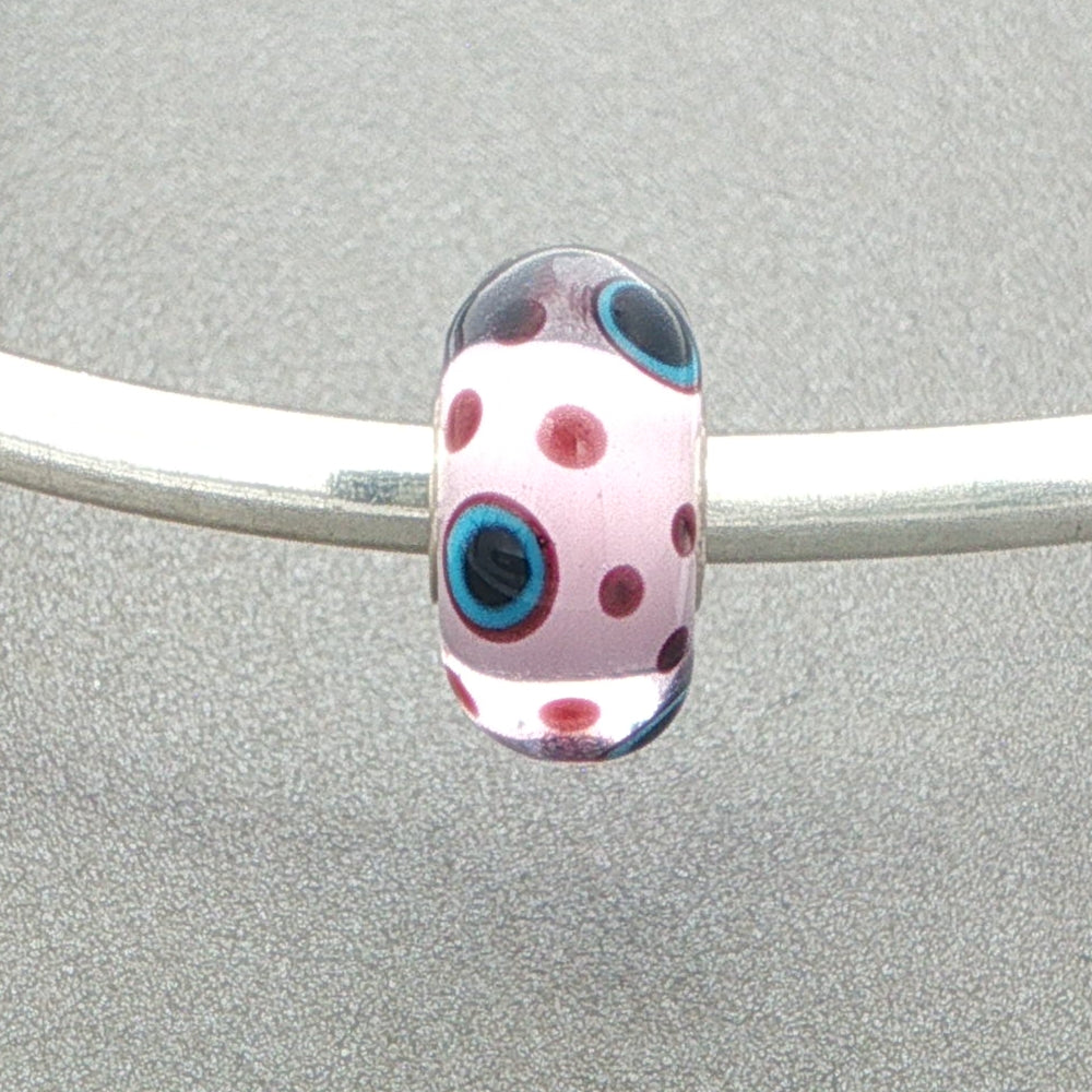 Polka dot murano glass bead with a pink core and burgundy dots with some with blue and black centres on a silver bangle against a grey background