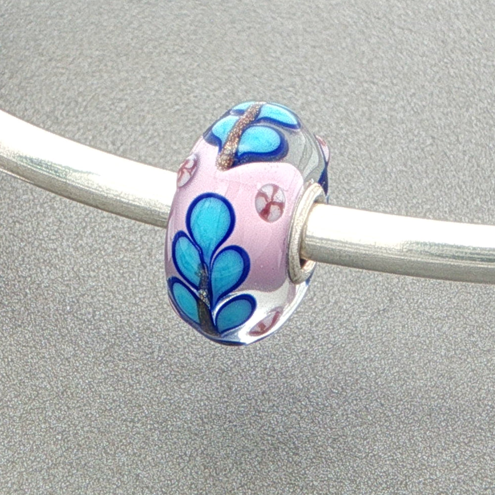 Glass bead with blue floral design on a silver bangle against a grey background