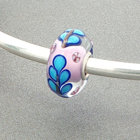 Glass bead with blue floral design on a silver bangle against a grey background