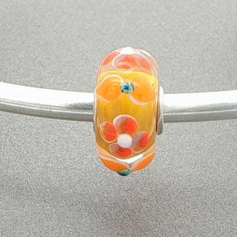Glass bead with an orange and yellow floral design on a silver bangle against a grey background