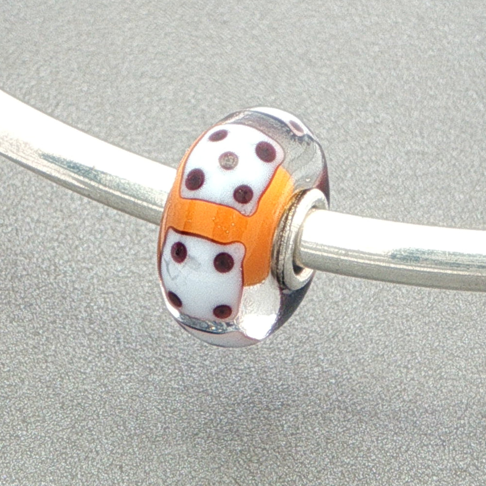 Glass bead with white dice design and orange core on a silver bangle against a grey background