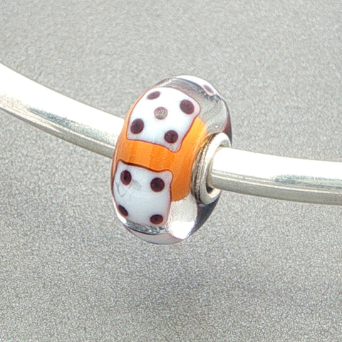 Glass bead with white dice design and orange core on a silver bangle against a grey background